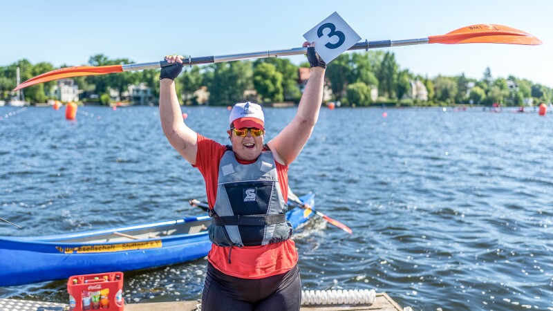 The photo shows kayakist Juliana Rößler after winning the kayaking race during the Special Olympics National Games Berlin 2022, holding her paddle above her head in jubilation.