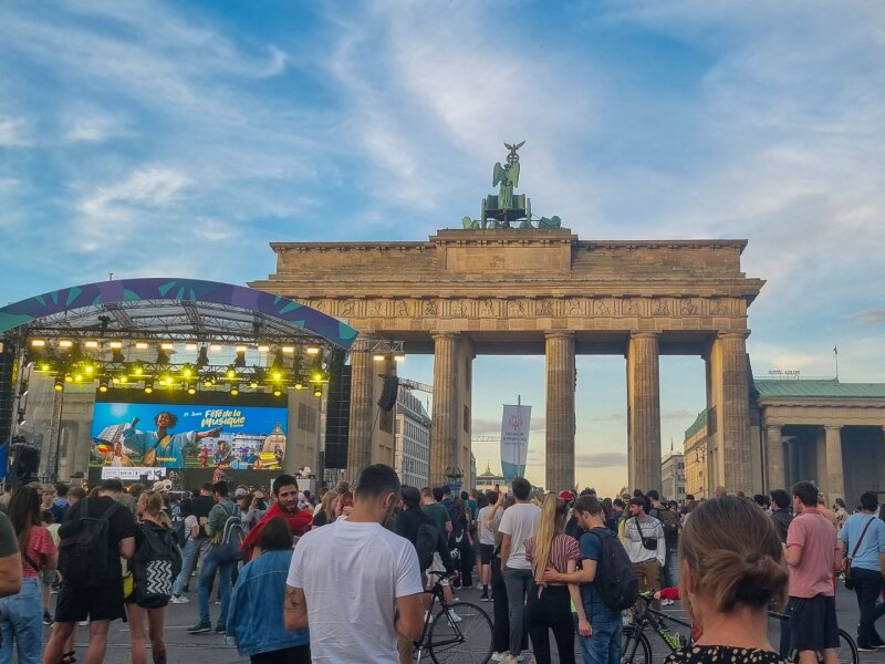 The photo shows a lively scene in front of the Brandenburg Gate. The stage in cooperation with the Fete de la Musique is set up directly in front of it and many people are milling around.