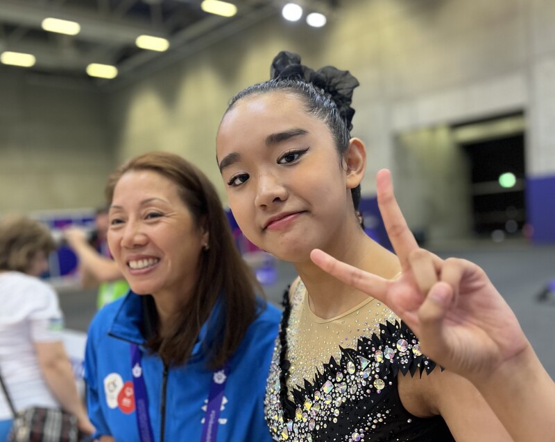 There are two people in the photo: A rhythmic gymnastics athlete is showing "peace" and the woman next to her is laughing happily.