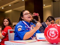 The photo shows a Special Olympics ambassador with a microphone. She is speaking and everyone is listening to her. In front of her is a soccer ball.