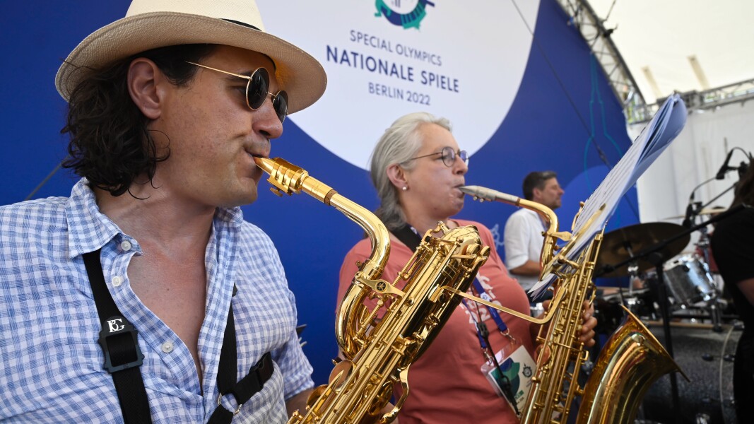The photo shows two men and one woman on a stage. In the foreground a man and a woman are playing a piece with their saxophone. In the background you can see the second man playing his drums.