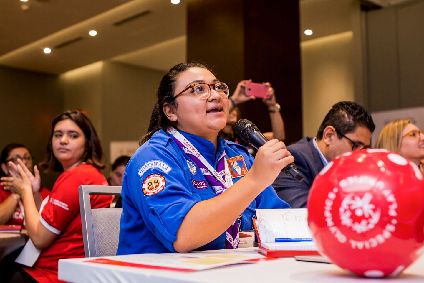 The photo shows a Special Olympics ambassador with a microphone. She is speaking and everyone is listening to her. In front of her is a soccer ball.