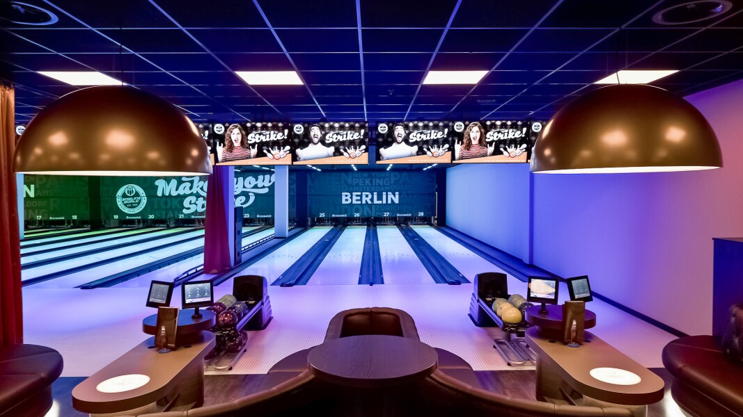 Das Foto zeigt einen Sitzbereich mit Monitoren und mehrere Bowlingbahnen im Hintergrund in einem Berliner Bowlingcenter.