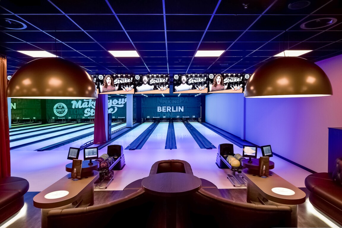 Das Foto zeigt einen Sitzbereich mit Monitoren und mehrere Bowlingbahnen im Hintergrund in einem Berliner Bowlingcenter.