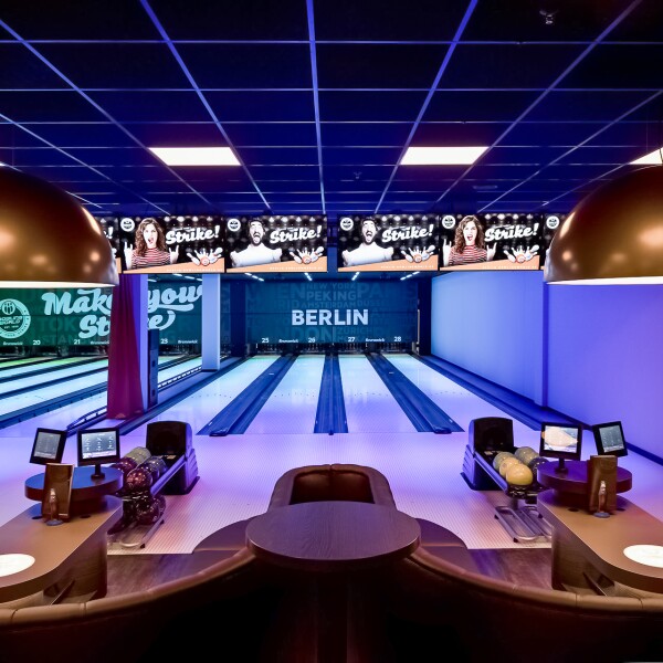 Das Foto zeigt einen Sitzbereich mit Monitoren und mehrere Bowlingbahnen im Hintergrund in einem Berliner Bowlingcenter.