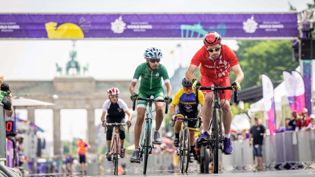 Das Foto zeigt mehrere Radsportler, die einander im Straßenrennen mit dem Brandenburger Tor einander verfolgen. Ganz vorn fährt ein Sportler mit rotem Trikot und kräftigem Antritt, verfolgt von einem Radfahrer in grüner Kleidung.