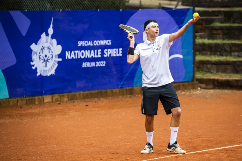 The photo shows a scene during the tennis competition at the Special Olympics National Games Berlin 2022. The player in the foreground is serving and in the background you can see a banner advertising the games. These banners were upcycled for products in our webshop.