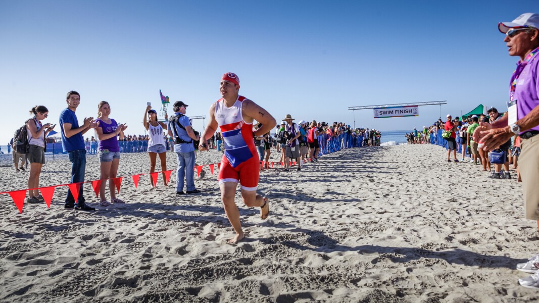 Das Foto zeigt einen Athleten beim Triathlon. Er steigt aus dem Wasser und rennt in die Wechselzone. Die Zuschauer*innen jubeln ihm zu.