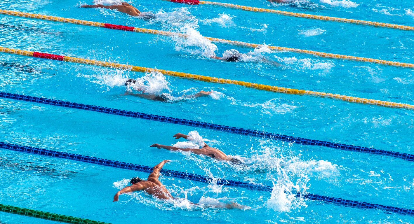 Das Foto zeigt mehrere Schwimmbahnen in einer Schwimmhalle und Menschen die schwimmen.