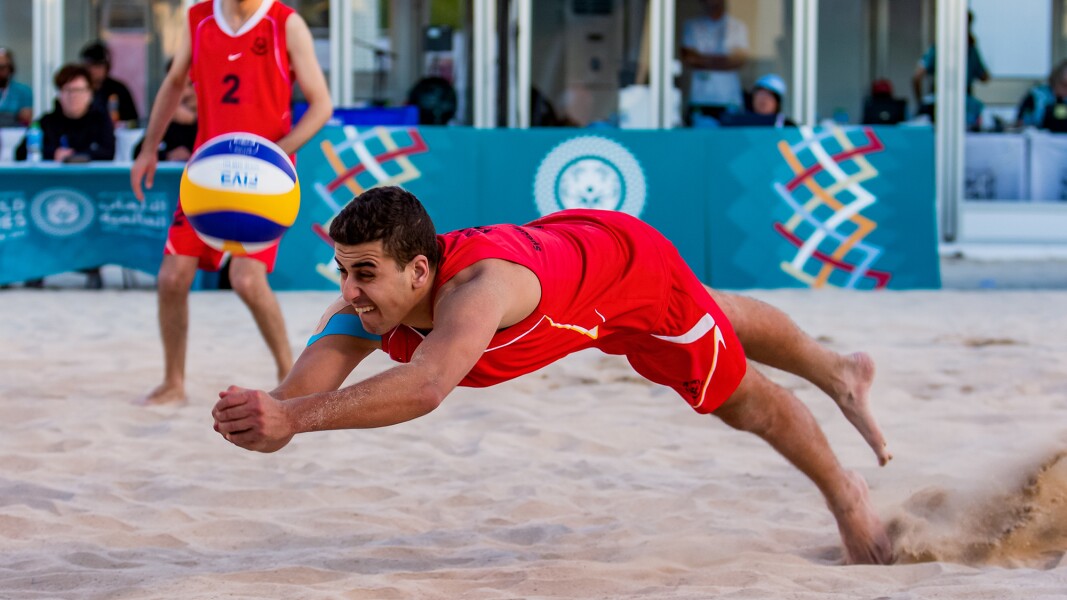 Auf dem Foto ist ein Beachvolleyballspieler zu sehen, der gerade spektakulär nach dem Ball springt.