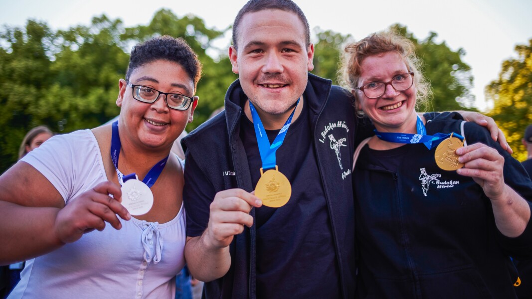 Auf dem Foto präsentieren stolz zwei Athletinnen und ein Athlet ihre Mediallen, zweimal Gold und einmal Silber. Sie lächeln und freuen sich.