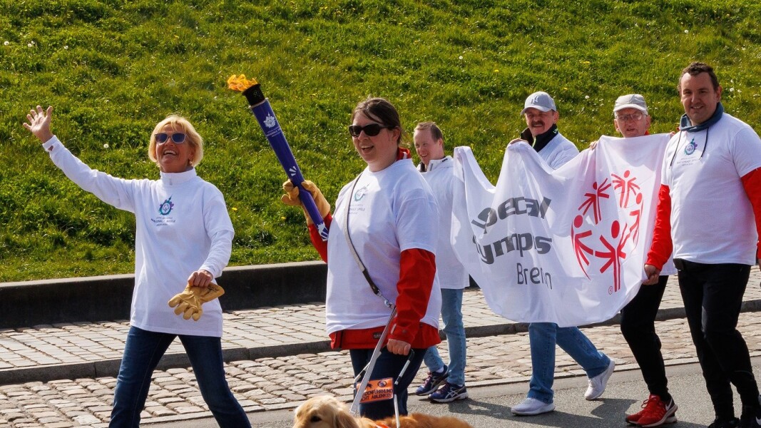 Das Foto zeigt eine Szene des Bremer Fackellaufes auf dem symbolischen Weg zu den Special Olympics Nationalen Spielen Berlin 2022. Es sind insgesamt 6 Menschen und ein Begleithund zu sehen. Die glücklich lächelnden Personen tragen eine Special Olympics Fahne und eine Athletin trägt die Fackel.