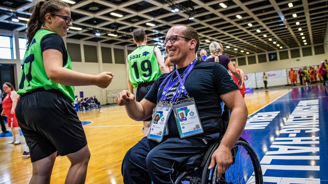Auf dem Foto ist die Halle im Basketball. Im Vordergrund ist eine Athletin, die gerade mit einem Mann im Rollstuhl spricht. Beide lachen fröhlich.