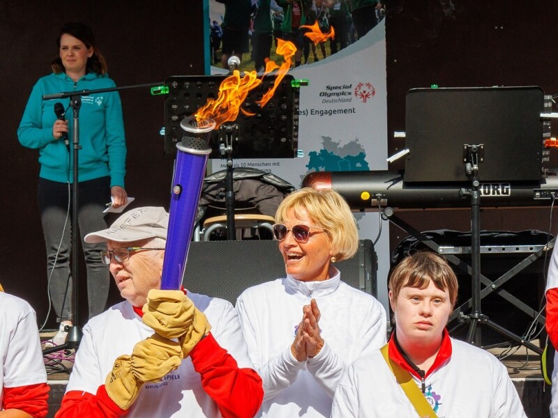Das Foto zeigt eine Gruppe Menschen, die am Bremer Fackellauf auf dem symbolischen Weg zu den Nationalen Spielen Berlin 2022 teilnehmen. Ein Mann trägt die Fackel und alle klatschen in die Hände. Hinter der Gruppe ist eine Bühne aufgebaut.
