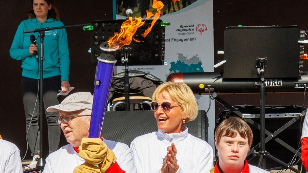 Das Foto zeigt eine Gruppe Menschen, die am Bremer Fackellauf auf dem symbolischen Weg zu den Nationalen Spielen Berlin 2022 teilnehmen. Ein Mann trägt die Fackel und alle klatschen in die Hände. Hinter der Gruppe ist eine Bühne aufgebaut.