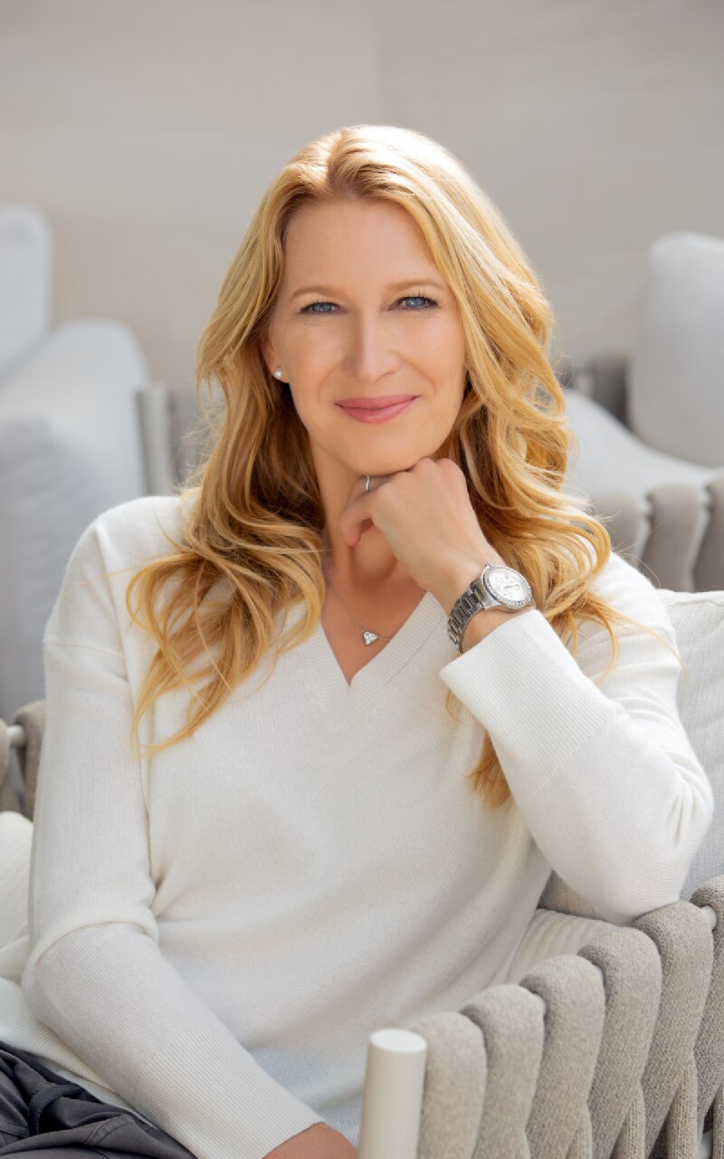 Auf dem Foto ist Steffi Graf, die deutsche Tennis-Legende, zu sehen. Sie trägt einen schlichten weißen Pullover und guckt freundlich in die Kamera.