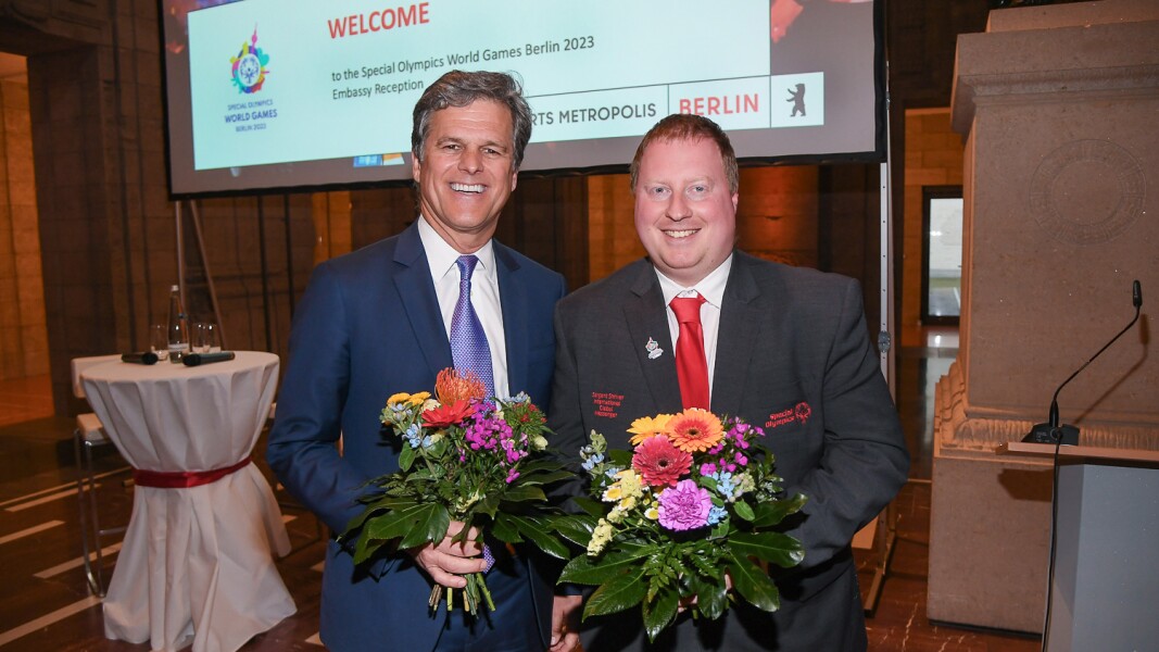 Das Foto zeigt Special Olympics International Chairman Timothy Shriver und Global Ambassador Ian Harper jeweils mit einem Blumenstraß in den Händen und einem Lächeln auf dem Gesicht.