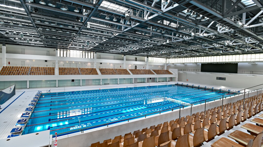 Das Foto zeigt ein Schwimmbecken, sowie links und rechts davon Zuschauerränge mit Holzstühlen.
