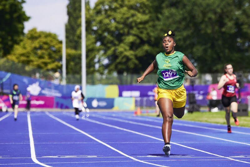 Four athletes running on the blue track while the Jamaican athlete Sheneel Williams in the yellow shorts and green jersey is approaching the finishing line.