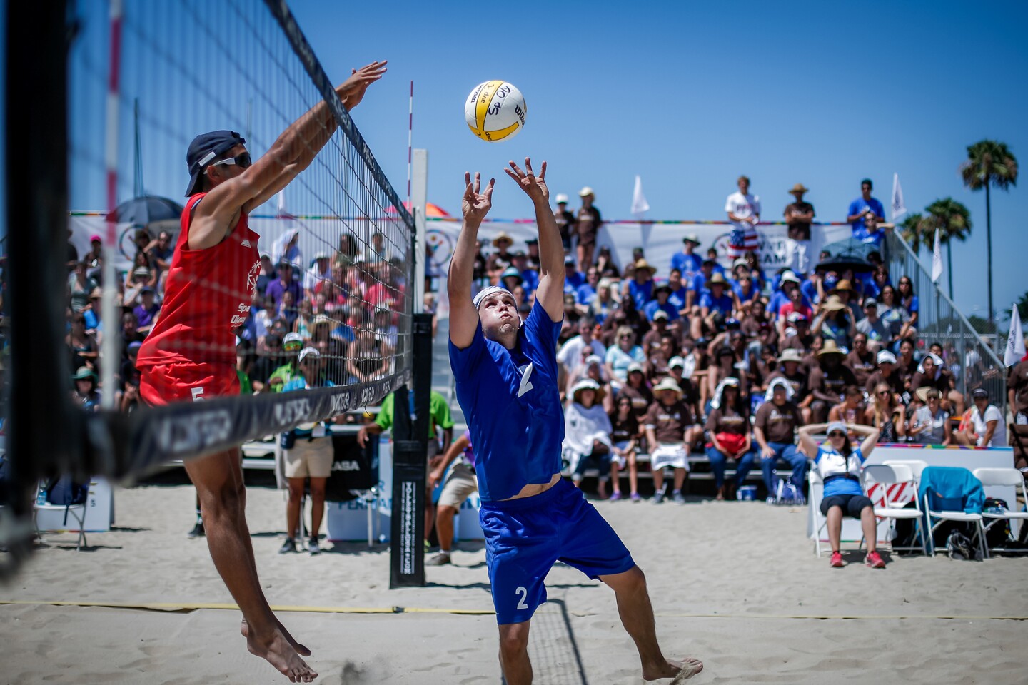 Das Bild zeigt eine Spielszene beim Beachvolleyball. Ein Spiel versucht am Netz zu blocken, während der Gegenspieler pritscht.