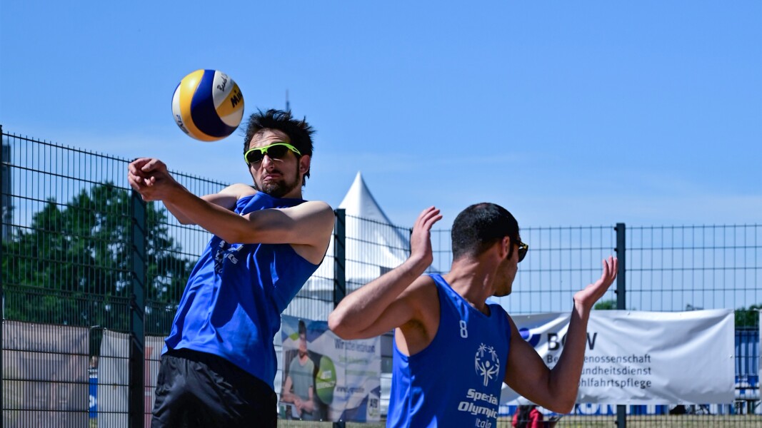Auf dem Foto sieht man zwei Athleten beim Beach-Volleyball. Der eine schlägt gerade den Bach-Volleyball.