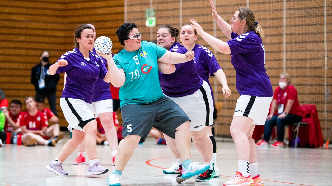 Auf dem Foto ist eine Gruppe Athletinnen zu sehen, eine Handballerin ist dabei den Handball zu werfen. Fünf Athletinnen der gegnerischen Mannschaft versuchen sie davon abzuhalten.