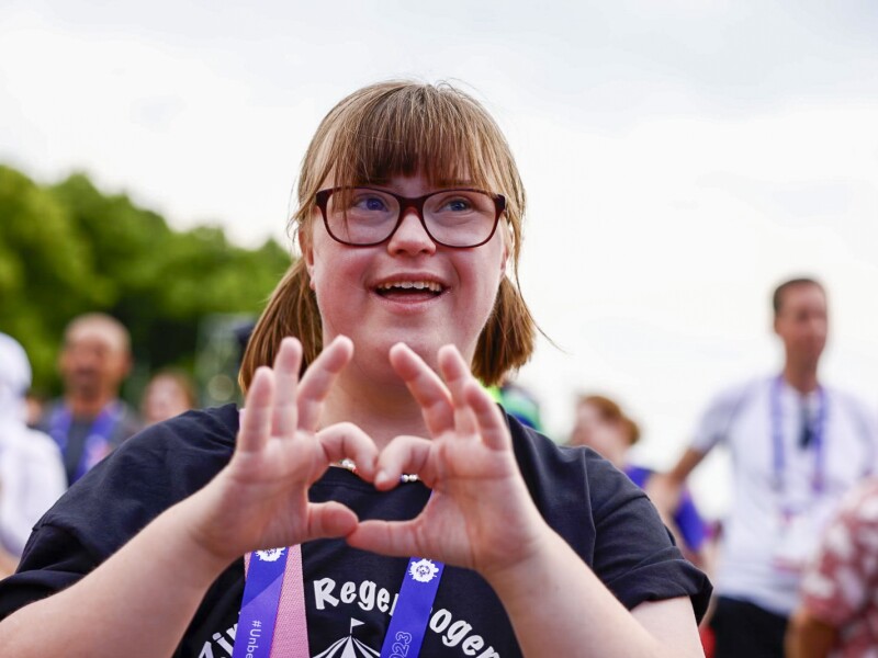 Das Foto zeigt ein Mädchen in einer Menschenmenge. Sie formt ein Herz mit ihren Händen in die Kamera und lacht.