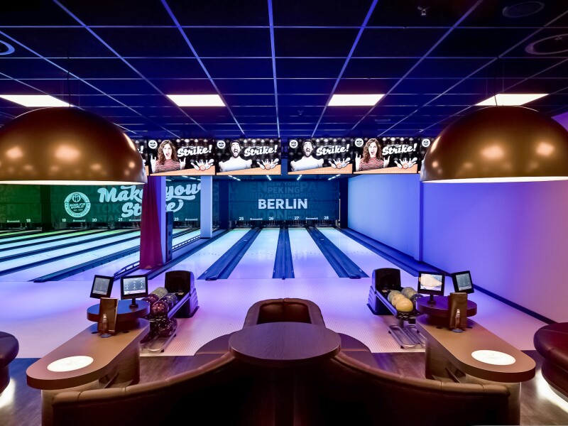 The photo shows a seating area with monitors and several bowling lanes in the background in a bowling center in Berlin.