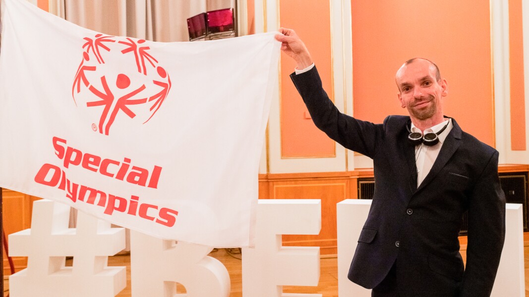 Auf dem Foto sieht man unser Gesicht der Nationalen Spiele Robert Herberg, der eine Special Olympics Flagge hält und in die Kamera lächelt.