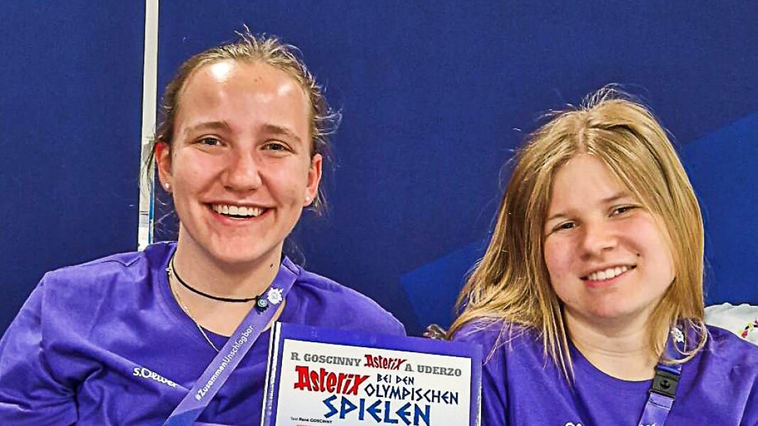 Das Bild zeigt die beiden Volunteers Clara und Luisa, wie sie am Stand der Unterlagenausgabe lächelnd eine Sonderausgabe von "Asterix at the Olympic Games" in die Höhe halten.