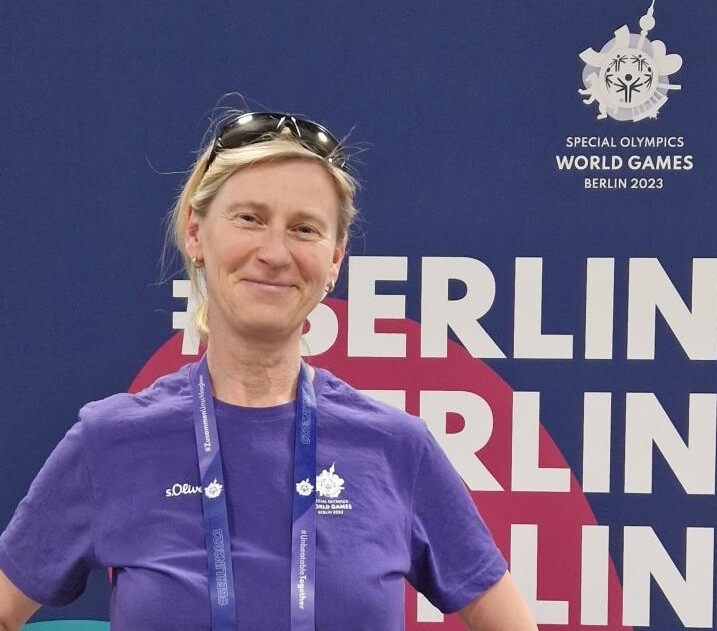 Das Bild zeigt die Volunteerin Ines stolz stehend vor dem Stand der Materialausgabe. Im Hintergrund stehen ganz viele gestapelte Kartons mit noch nicht ausgepackten Informationsbroschüren.