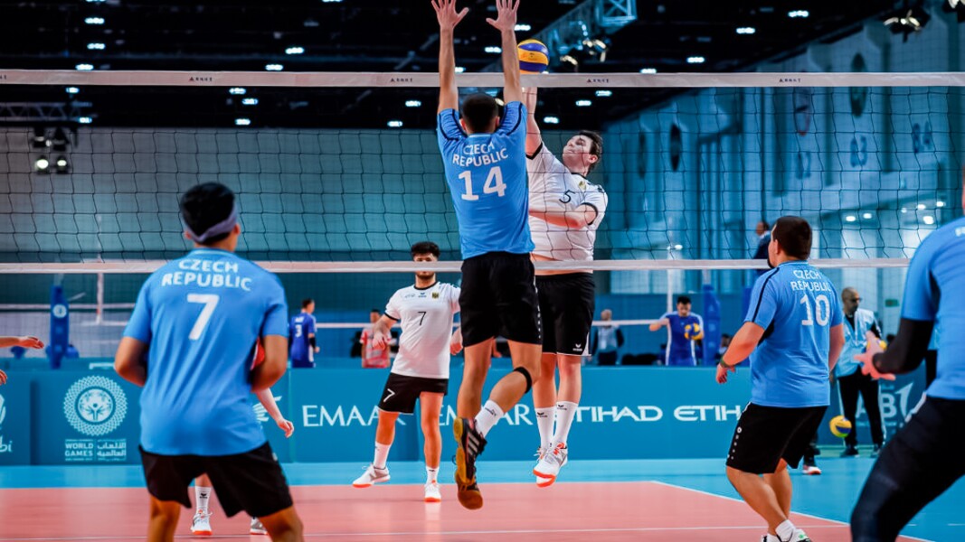 Das Foto zeigt zwei Mannschaften beim Volleyball. Zwei Spieler sind in Aktion am Netz. Ein Spieler versucht den Ball über das Netz zu spielen, sein Gegner hat beide Arme nach oben gestreckt, um den Ball abzuwehren.