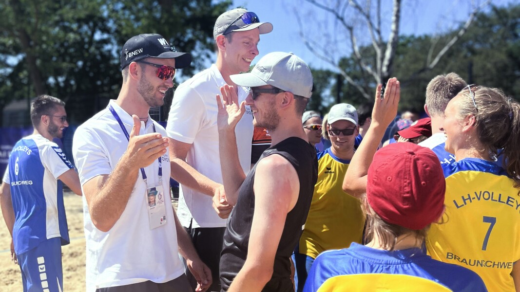 Das Foto zeigt die Beachvolleyball-Profis Nils Ehlers und Clemens Winkler, die mit vielen Athlet*innen beim Beachvolleyball während der Nationalen Spiele abklatschen und lächeln.