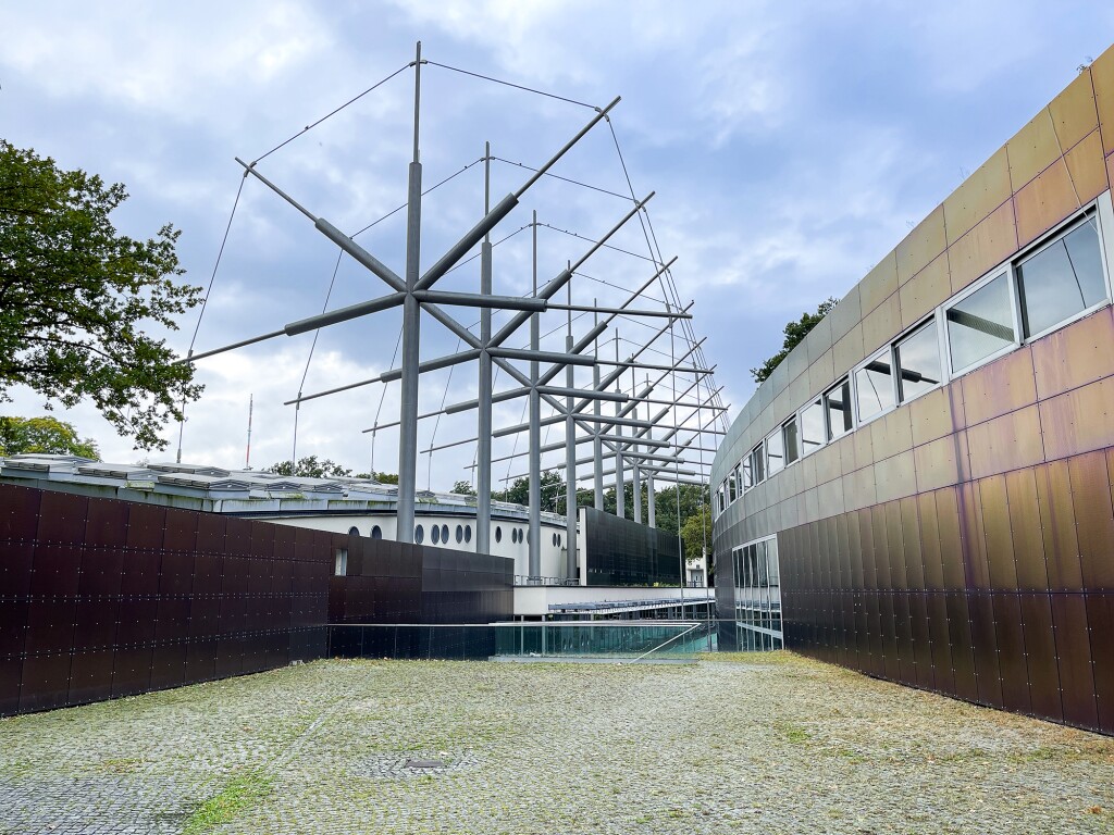 Das Foto zeigt ein weißes Gebäude im Hintergrund und ein Gebäude mit einer Metallfassade im Vordergrund. In der Mitte zwischen beiden Gebäuden ist eine Stahlkonstruktion zu sehen.