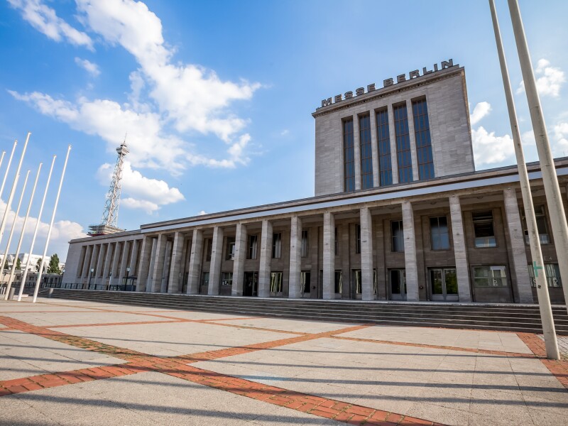 The photo shows a paved forecourt and a building with columns on which Messe Berlin stands.