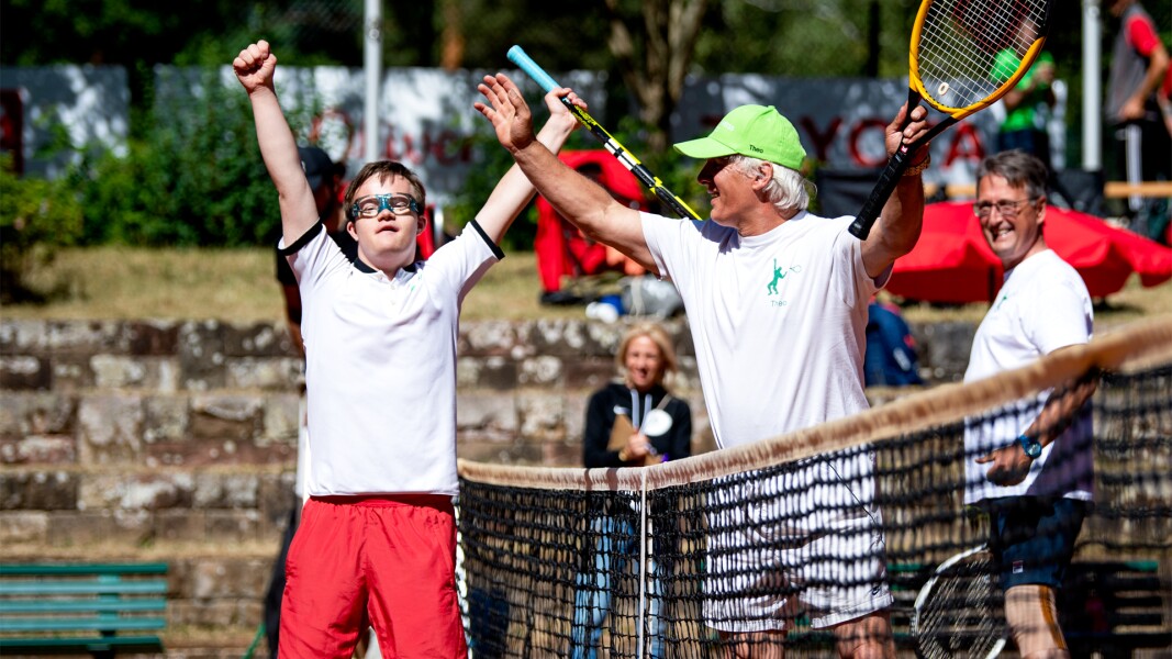Auf dem Foto sieht man einen Athlet auf dem Tennisplatz, der seine Arme nach oben streckt und sich freut. Auf der anderen Seite des Tennisnetzes steht auch noch ein Mann, der seine Arme und seinen Tennisschläger in die Luft streckt.