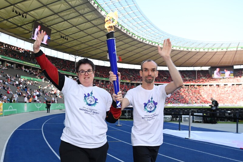 Robert Herberg, Gesicht der Special Olympics Nationalen Spiele Berlin 2022, und Athletin Juliana Rößler halten gemeinsam die Fackel von Special Olympics. Sie laufen auf der blauen Bahn im Berliner Olympiastadion.