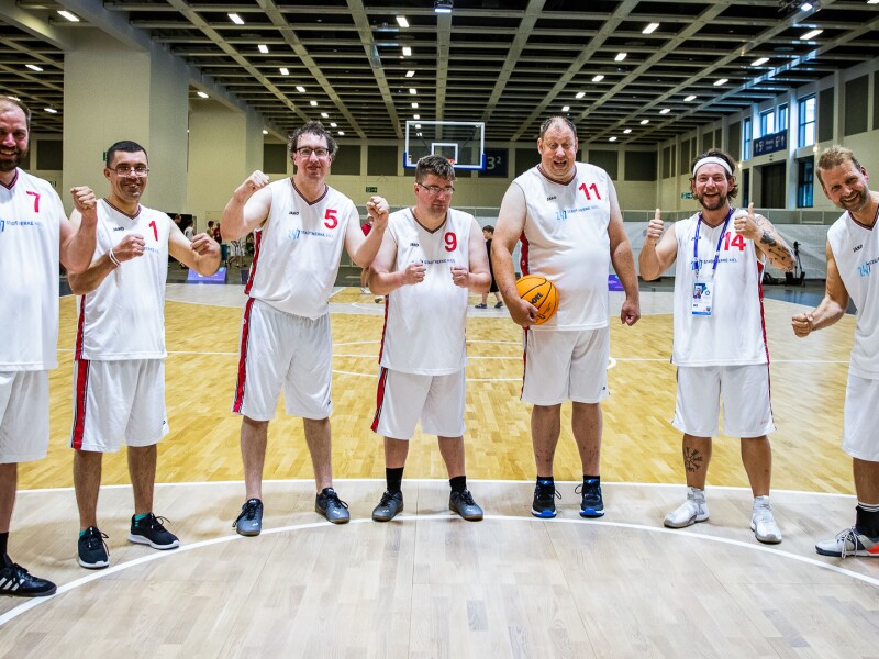 In the photo a basketball team is standing in a semicircle on the court. Everyone is cheering joyfully into the camera and one player is holding a basketball. It is not a game situation.