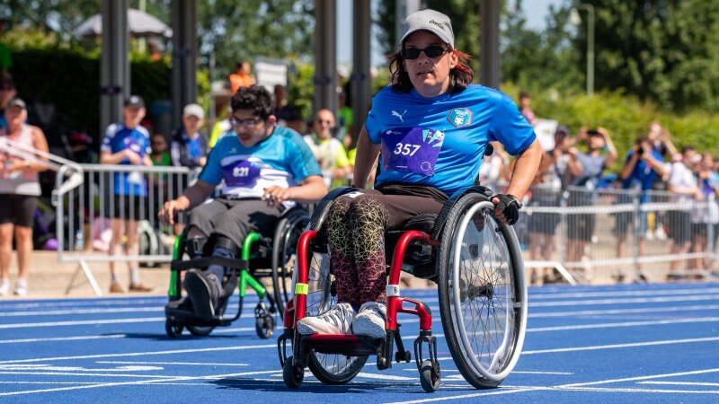 Auf dem Foto ist im Vordergrund eine Frau, die an den Leichtathletik-Wettbewerben im Rollstuhl teilnimmt. Sie trägt eine Mütze und eine Sonnenbrille und schaut in die Kamera. 