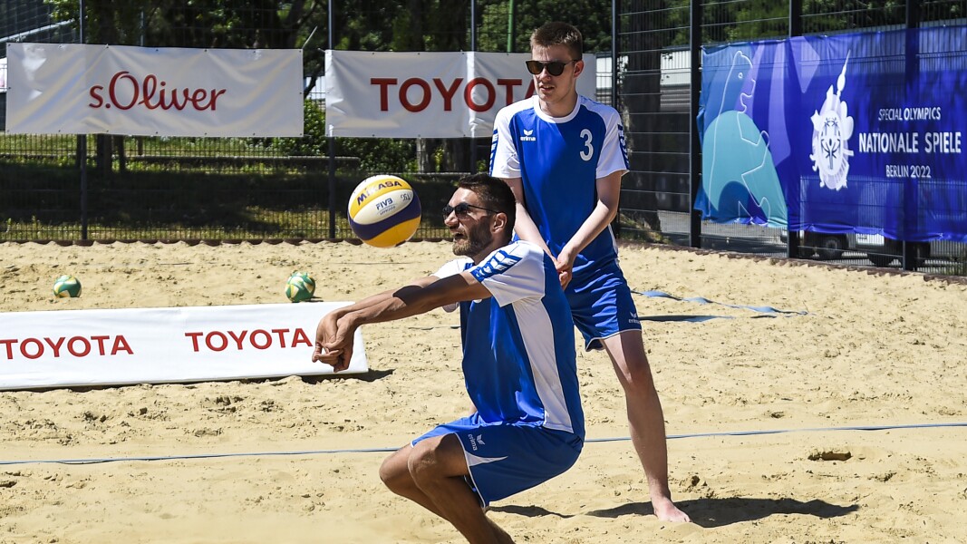 The photo shows two men playing beach volleyball. One is just accepting a ball and is squatting. The other is standing behind and looking at the ball. He is wearing sunglasses. In the background you can see many sponsor banners hanging on the fence.