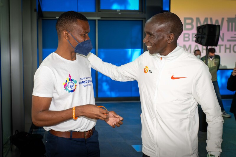 Das Foto zeigt Special Olympics International Global Messenger und Langstreckenläufer Nyasha Derera aus Simbabwe gemeinsam mit Olympiasieger und Vorbild Eliud Kipchoge aus Kenia. Kipchoge hat freundschaftlich seine Hand auf Dereras Schulter gelegt und beide lächeln sich gegenseitig zu. 