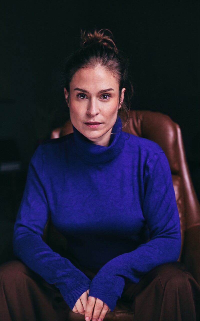 Das Foto zeigt die deutsche Schauspielerin Lea Marlen Woitack. Sie sitzt auf einem Sessel, blickt in die Kamera und trägt einen blauen Rollkragenpullover.