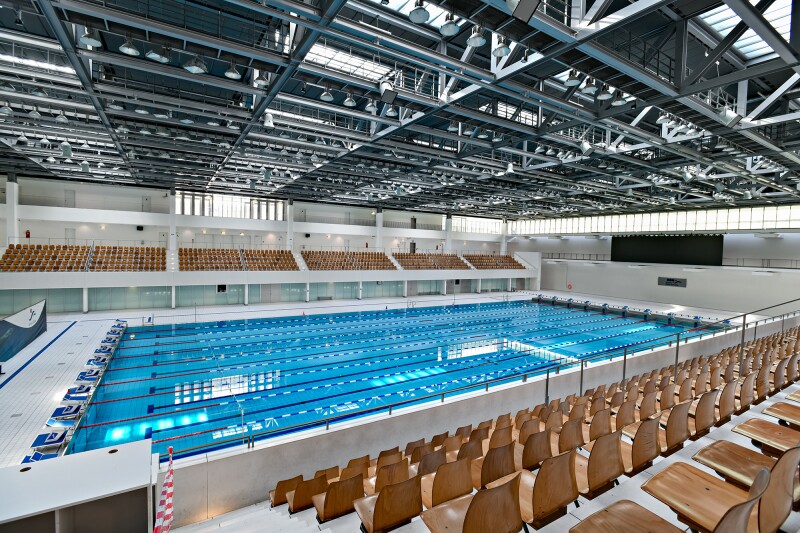 The photo shows a swimming pool, as well as spectator terraces with wooden chairs to the left and right of it.