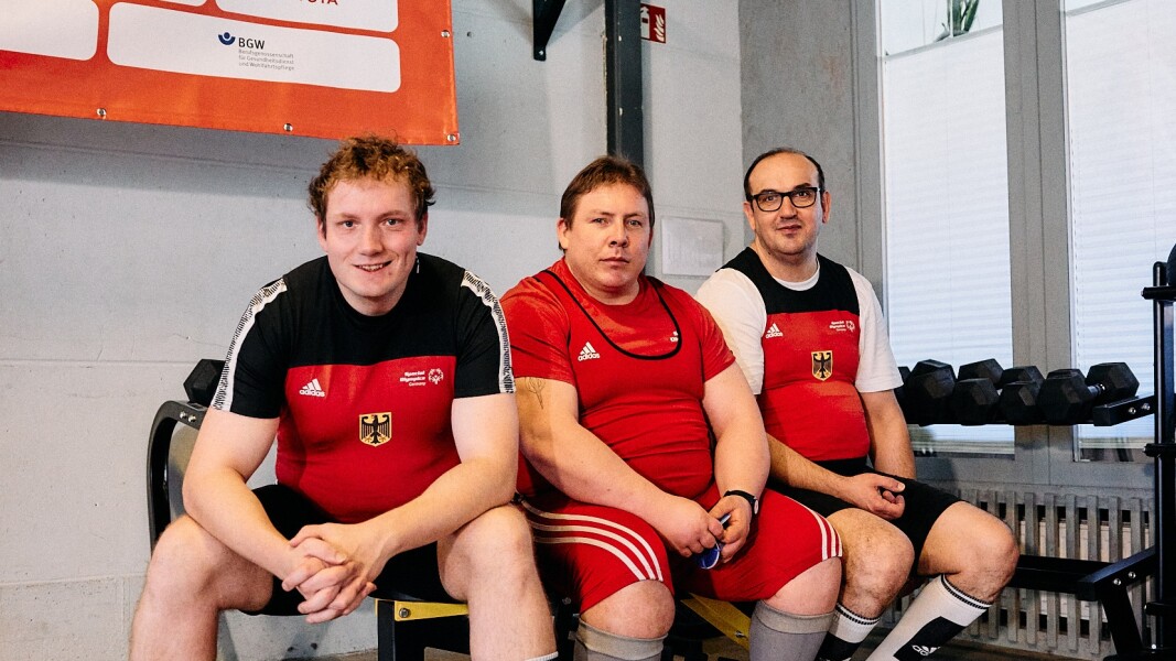 Auf dem Foto sitzen drei deutsche Athleten in ihren offiziellen Kraftdreikampf-Outfits auf einer Bank. Sie haben gerade ihre Sportart für Medienvertreter*innen demonstriert.