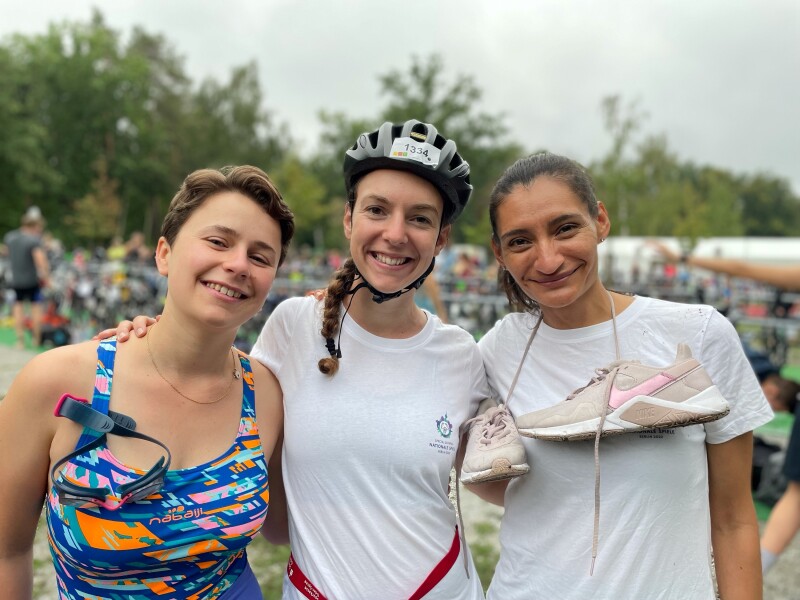 The photo shows three women participating in the BerlinMan Triahtlon. On the left in swimsuit and goggles, in the middle with a bike helmet and on the right with running shoes - symbolic for the three disciplines of the triathlon. All three smile relaxed into the camera.