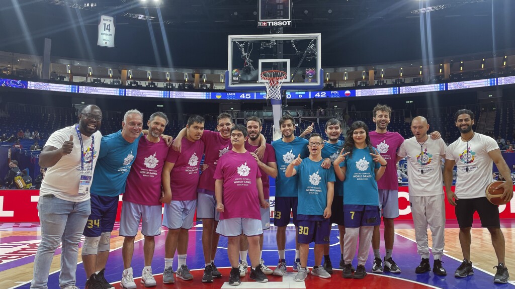 Das Foto ist ein Gruppenbild der Spieler*innen von Mina Basket auf dem Main Court der Basketball Europameisterschaft in Berlin, wo sie ein Halbzeit-Show-Match spielen durften. Das Unified Basketball Team liegt sich in den Armen und alle lächeln freudig in Richtung Kamera.