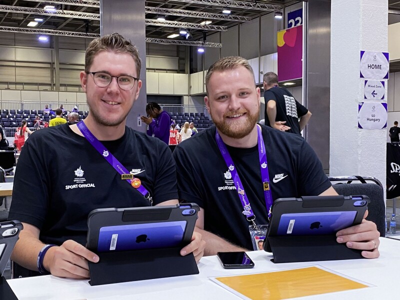 Das Foto zeigt die beiden Unified Observer Felix Beiser und Kilian Wich bei ihrer Arbeit beim Basketball. Beide halten ein Tablet in der Hand und lächeln freundlich.
