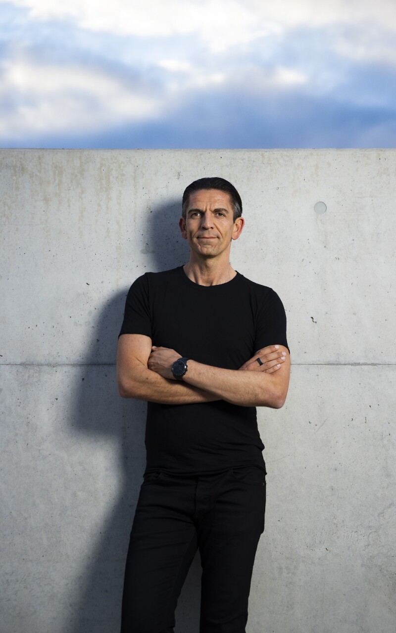 Auf dem Foto steht der deutsche Fußball-Schiedsrichter Deniz Aytekin mit verschränkten Armen und einem schlichten schwarzen Outfit vor einer grauen Mauer. Er blickt freundlich in Richtung Kamera.