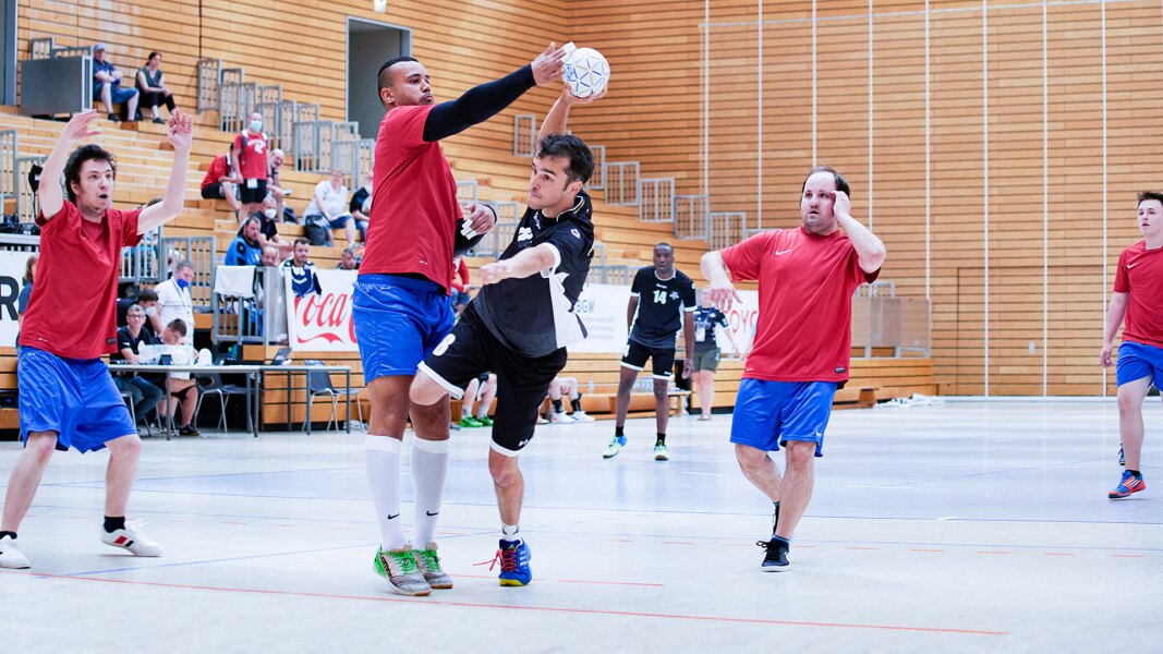 Das Foto zeigt Athleten bei einem Handballspiel in der Halle. Ein Spieler macht gerade einen Sprungwurf. Ein gegnerischer Spieler versucht ihn zu blocken. Im Hintergrund sind noch vier weitere Spieler zu sehen.