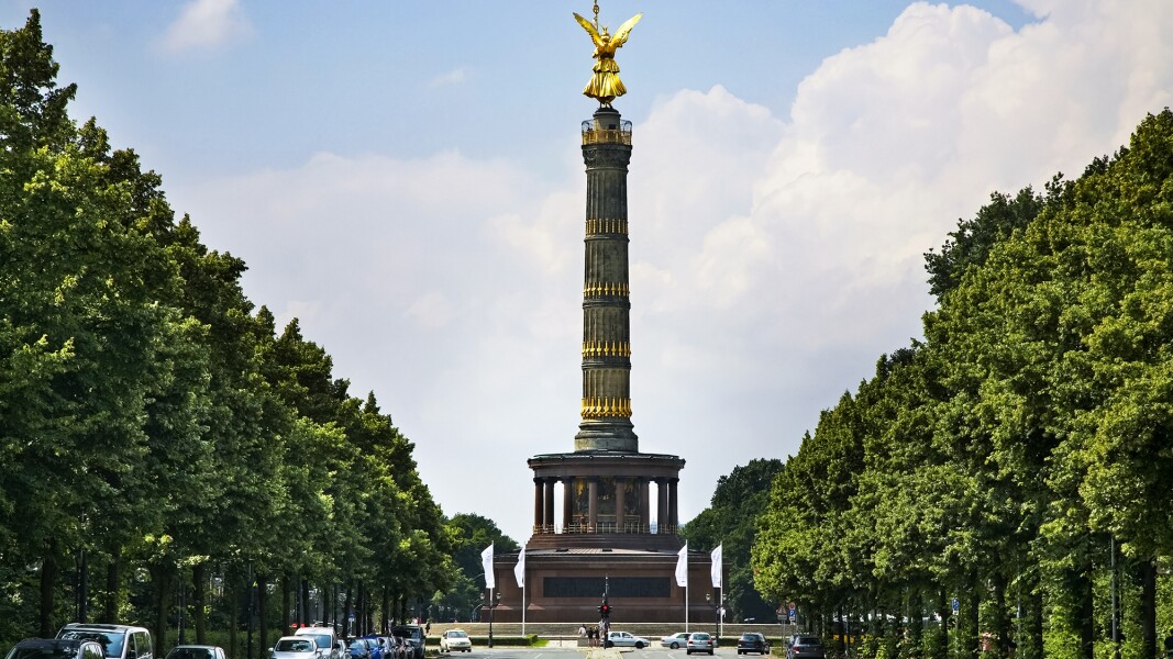 The photo shows a street with parked cars and in the center the Victory Column in Berlin.